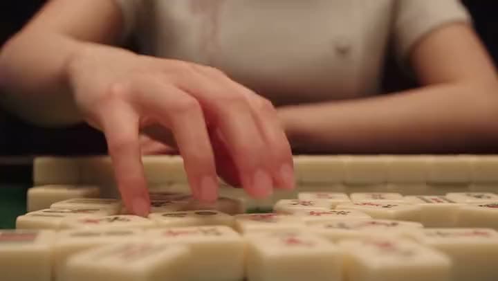 An AI video of a close-up of a woman's hand reaching over rows of mahjong tiles on a table.