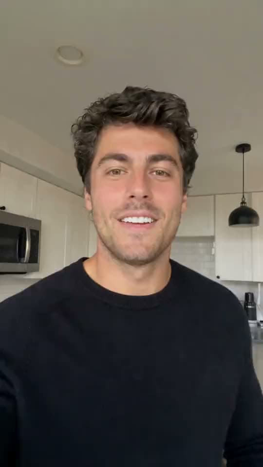 A vertical video of a smiling man with curly dark hair in a black sweater talking to his phone camera in a kitchen.