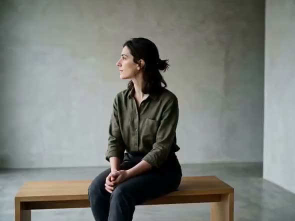 An AI video of a woman in an olive shirt sitting on a wooden bench in a bare concrete room, gazing off to the side.