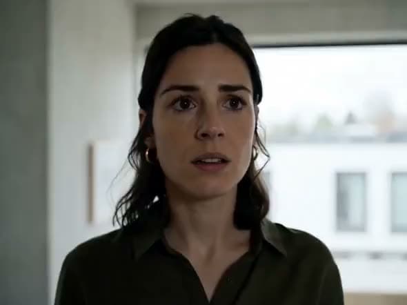 An AI video of a dark-haired woman in an olive shirt looking alarmed in a pale concrete room with a large window.