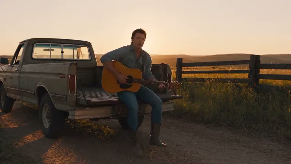 A man playing an acoustic guitar beside a pickup truck at sunset.