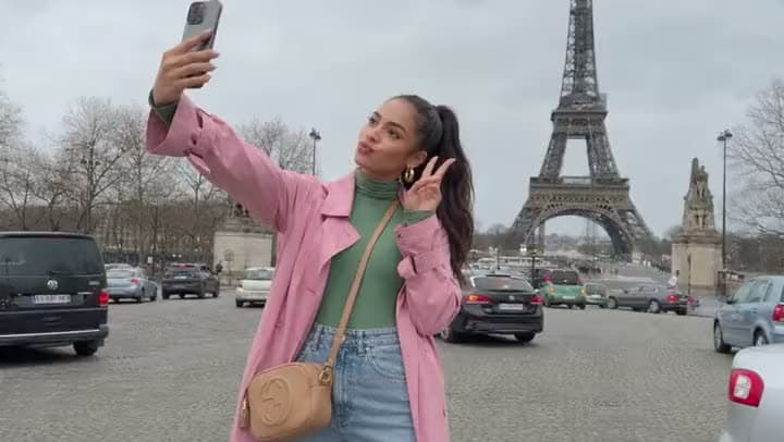 An AI video of a woman in a pink coat blowing a kiss while taking a selfie in front of the Eiffel Tower.