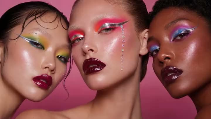 An AI video of three models with bold graphic eyeliner, colorful eyeshadow and dark glossy lips against a pink backdrop.