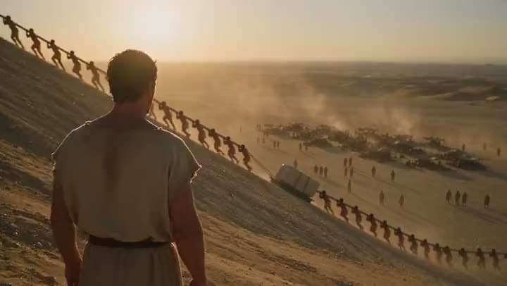 An AI video of a man in a tunic watching lines of workers haul a stone block up a sand ramp in the desert at sunset.
