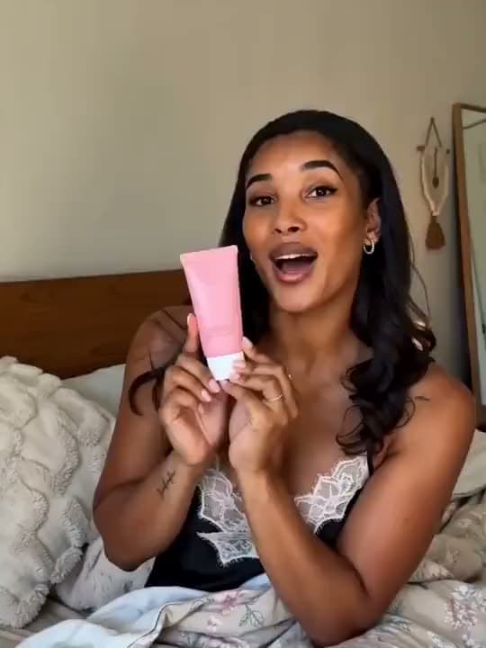 A vertical AI video of a woman in bed holding up a pink tube of skincare and talking to the camera in a UGC-style ad.