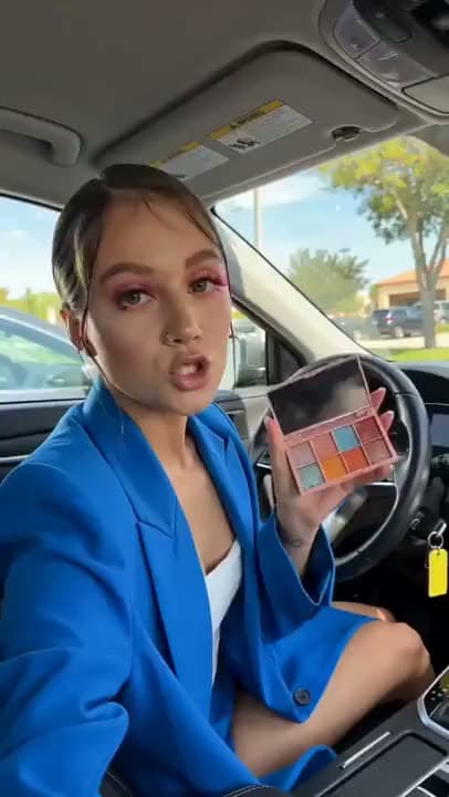 A vertical AI video of a woman in a blue blazer sitting in a parked car, holding up an eyeshadow palette as she talks to camera.