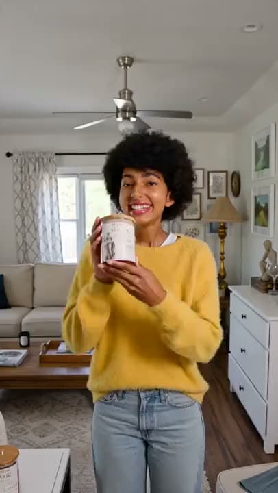 A vertical AI video of a smiling woman in a yellow sweater holding up a candle tin to the camera in a bright living room.
