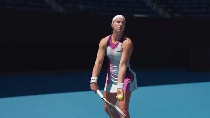 An AI video of a female tennis player in a pink and white dress bouncing the ball before a serve on a blue court.
