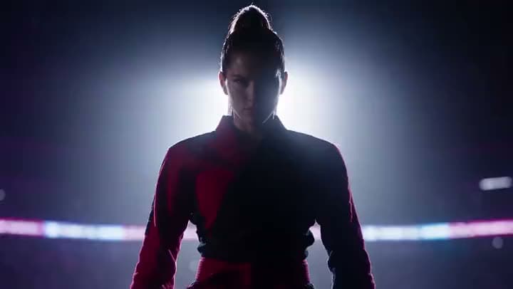 An AI video of a woman in a red and black jacket standing backlit by a bright spotlight in a dark fight arena.